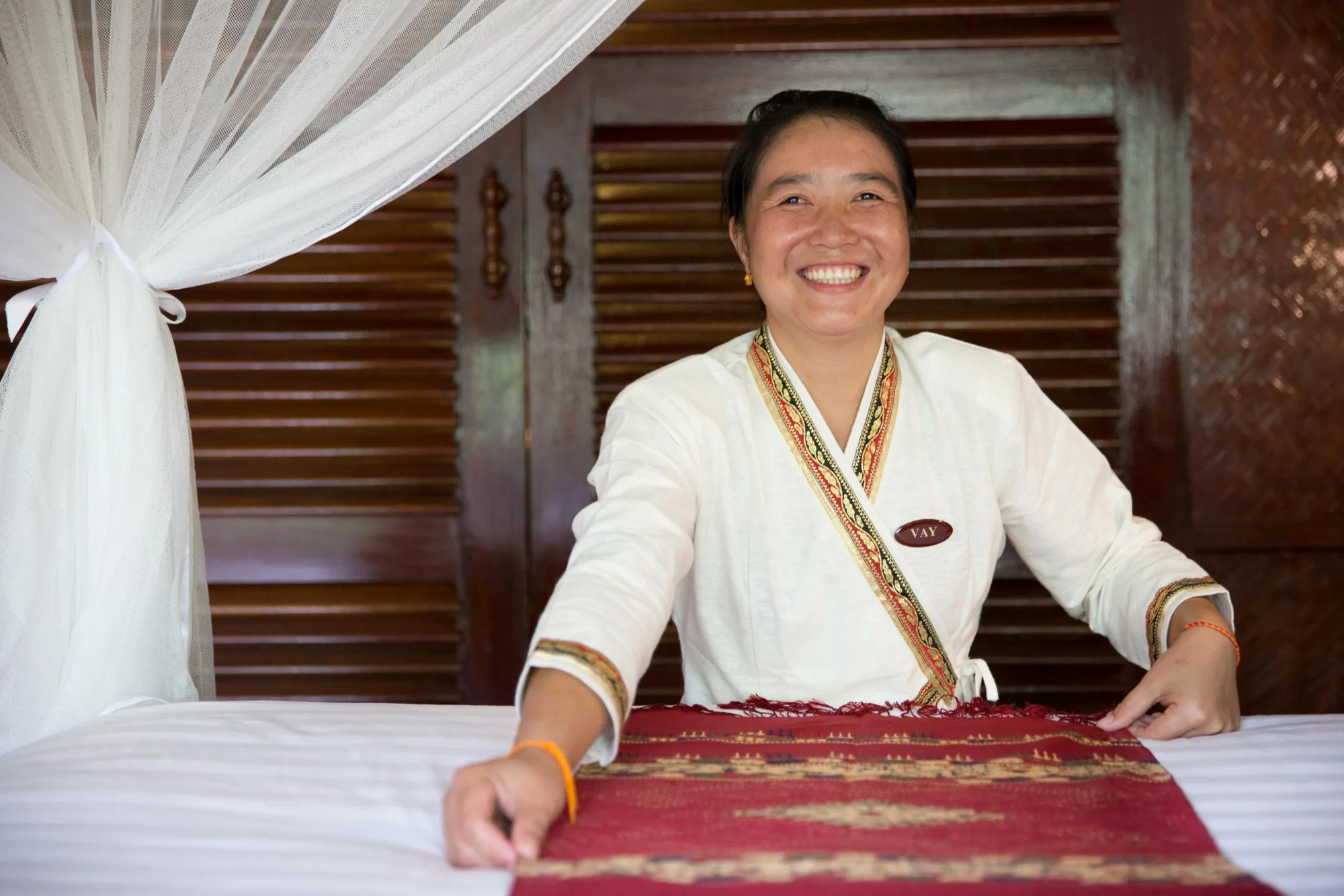 A smiling lodge staff member in traditional dress preparing a guest bed with a woven cotton throw