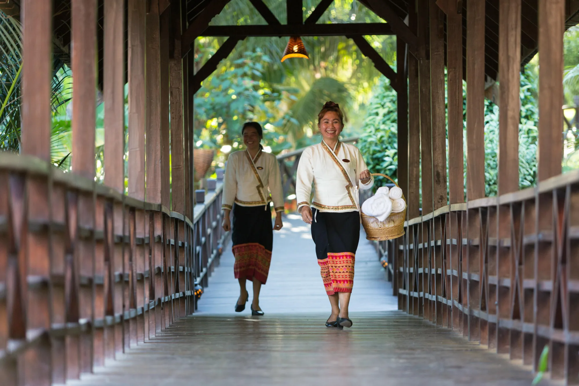 Two staff members in traditional Lao dress crossing a covered wooden walkway, one carrying a basket of fresh towels