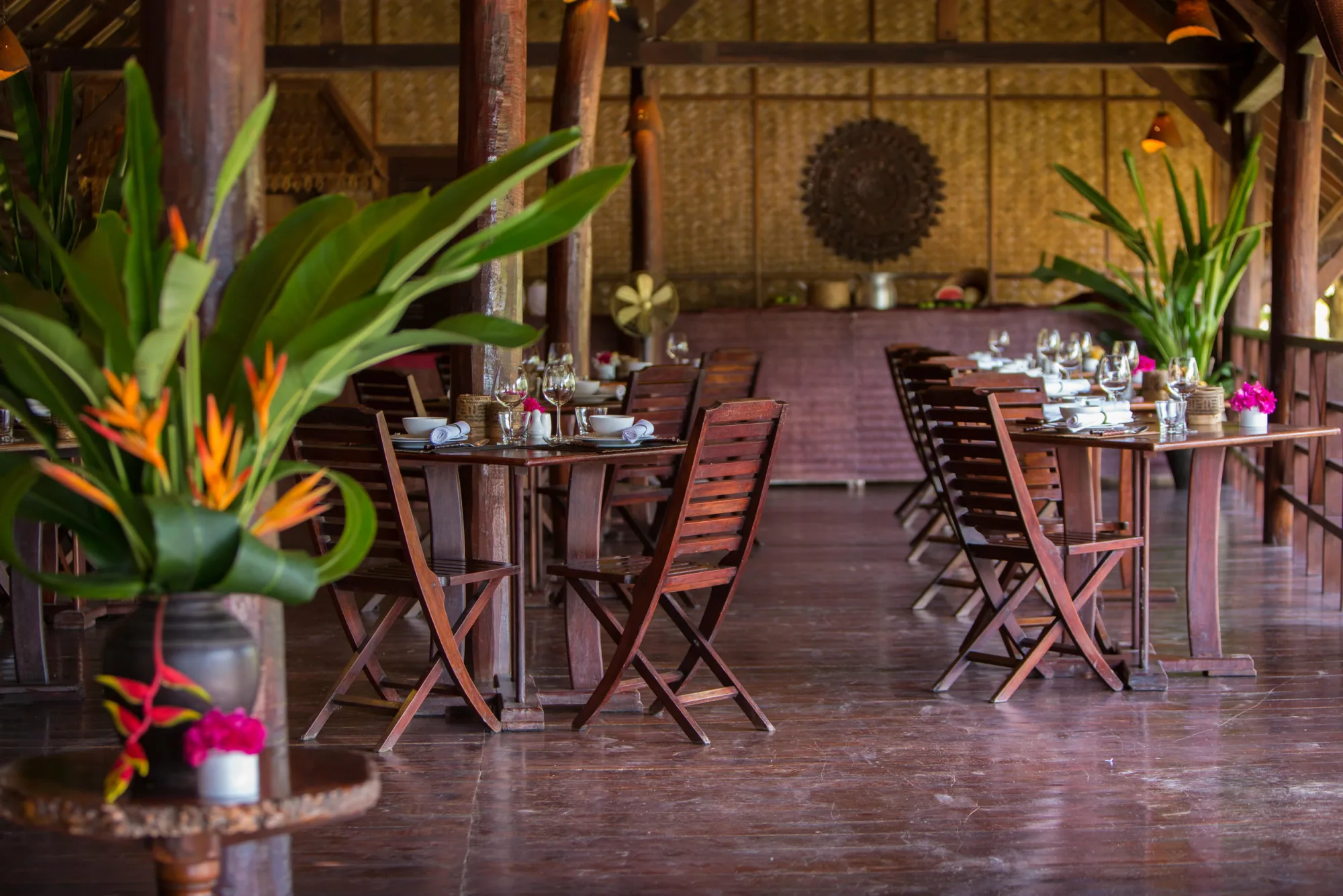 Riverside dining room under a high thatched roof, tables set with tropical flower arrangements
