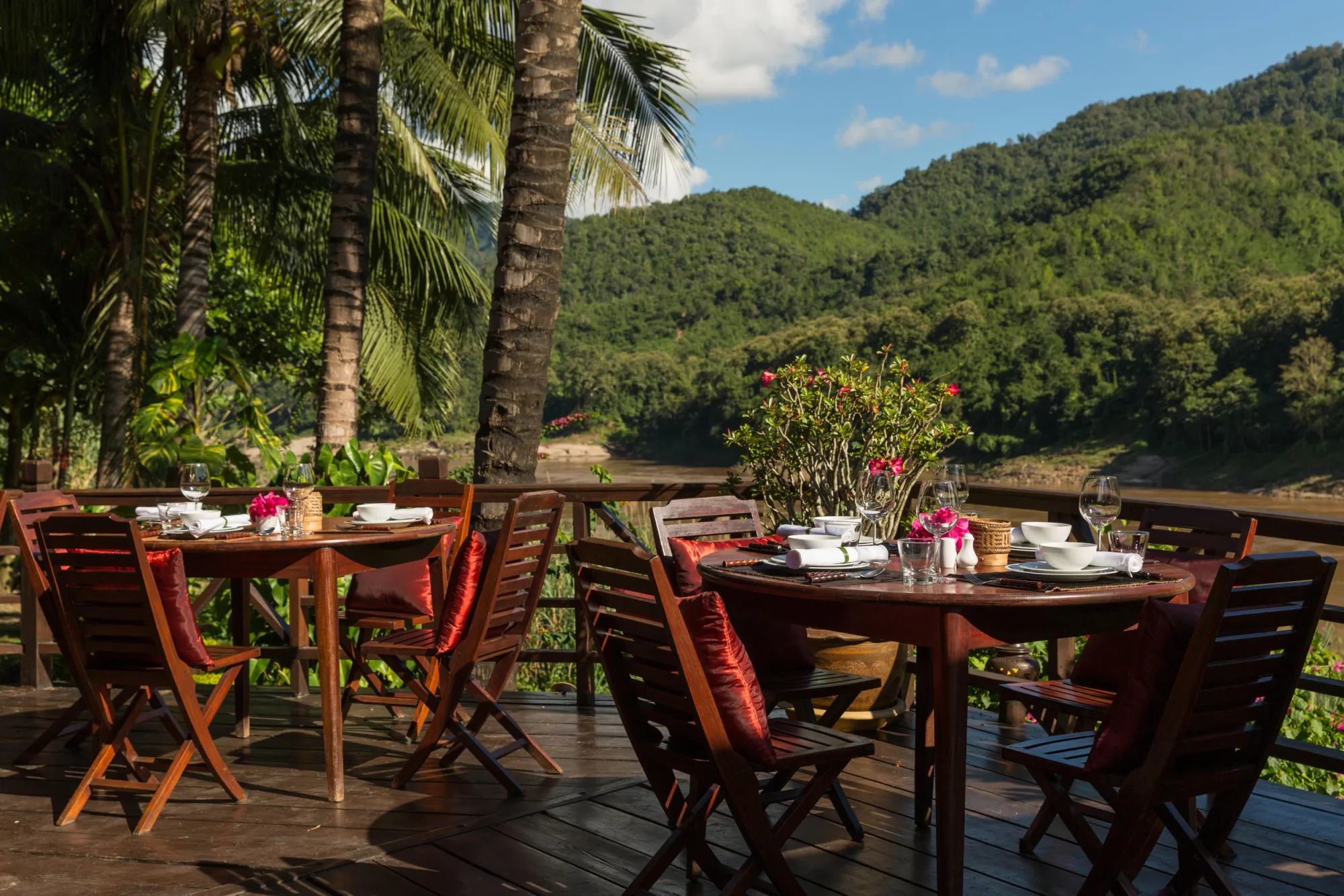 Round tables set for dining on the terrace among palm trees, with the Mekong and forested hills beyond