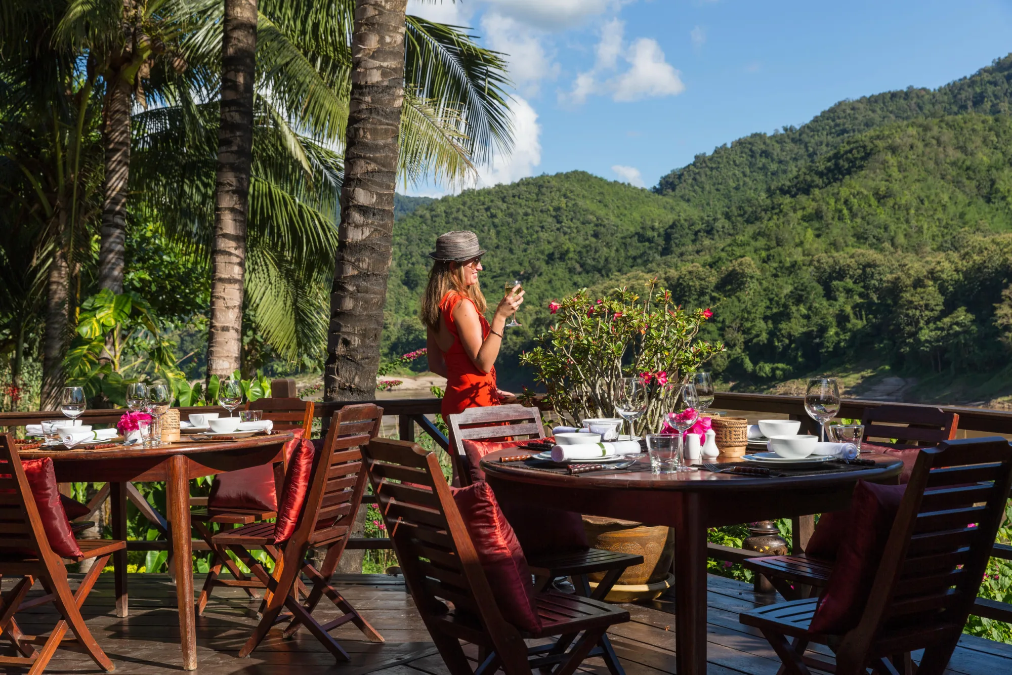 A guest holding a glass of wine on the terrace, with palm trees and the Mekong behind her