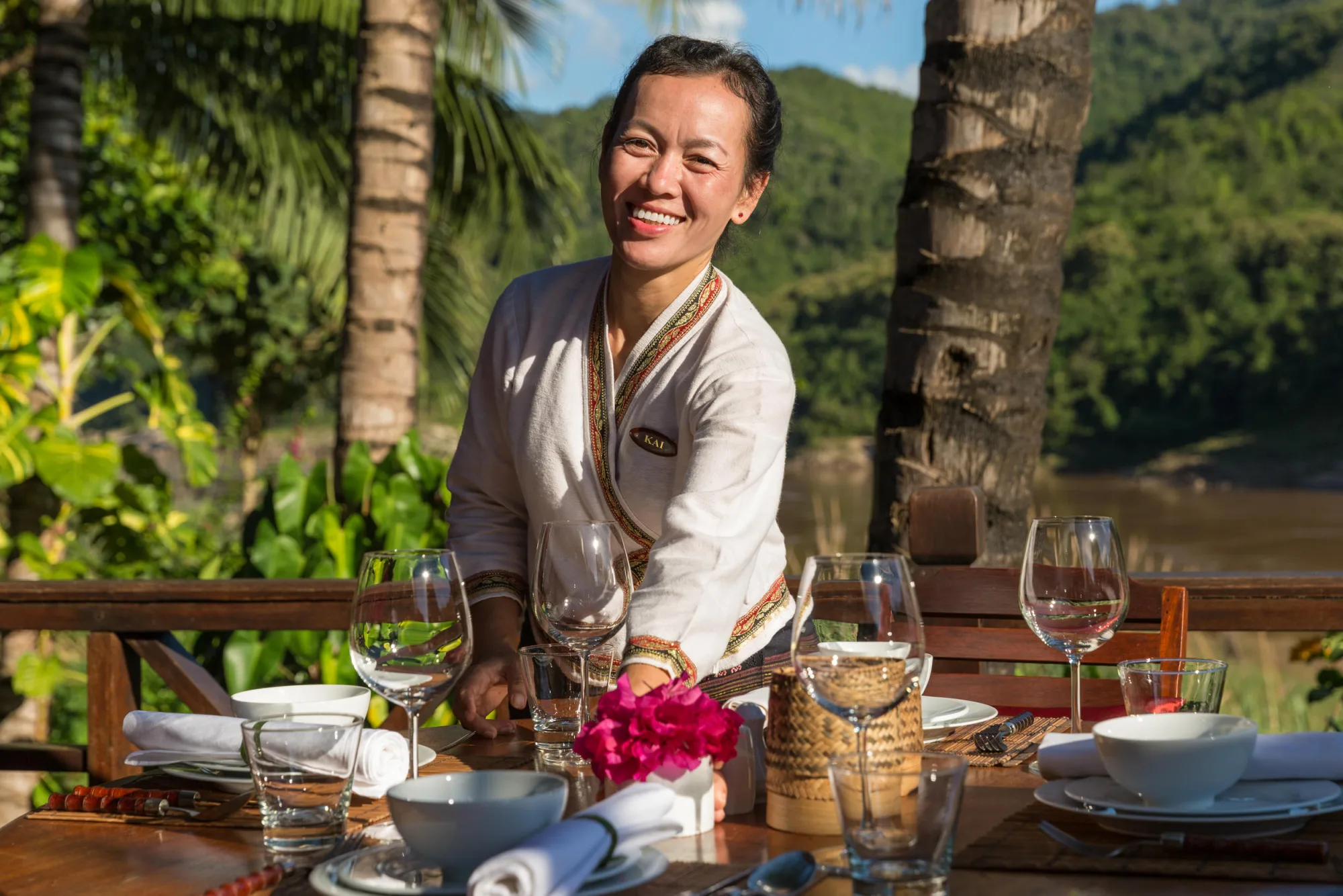 A smiling staff member setting a terrace table, with the Mekong and hills behind her