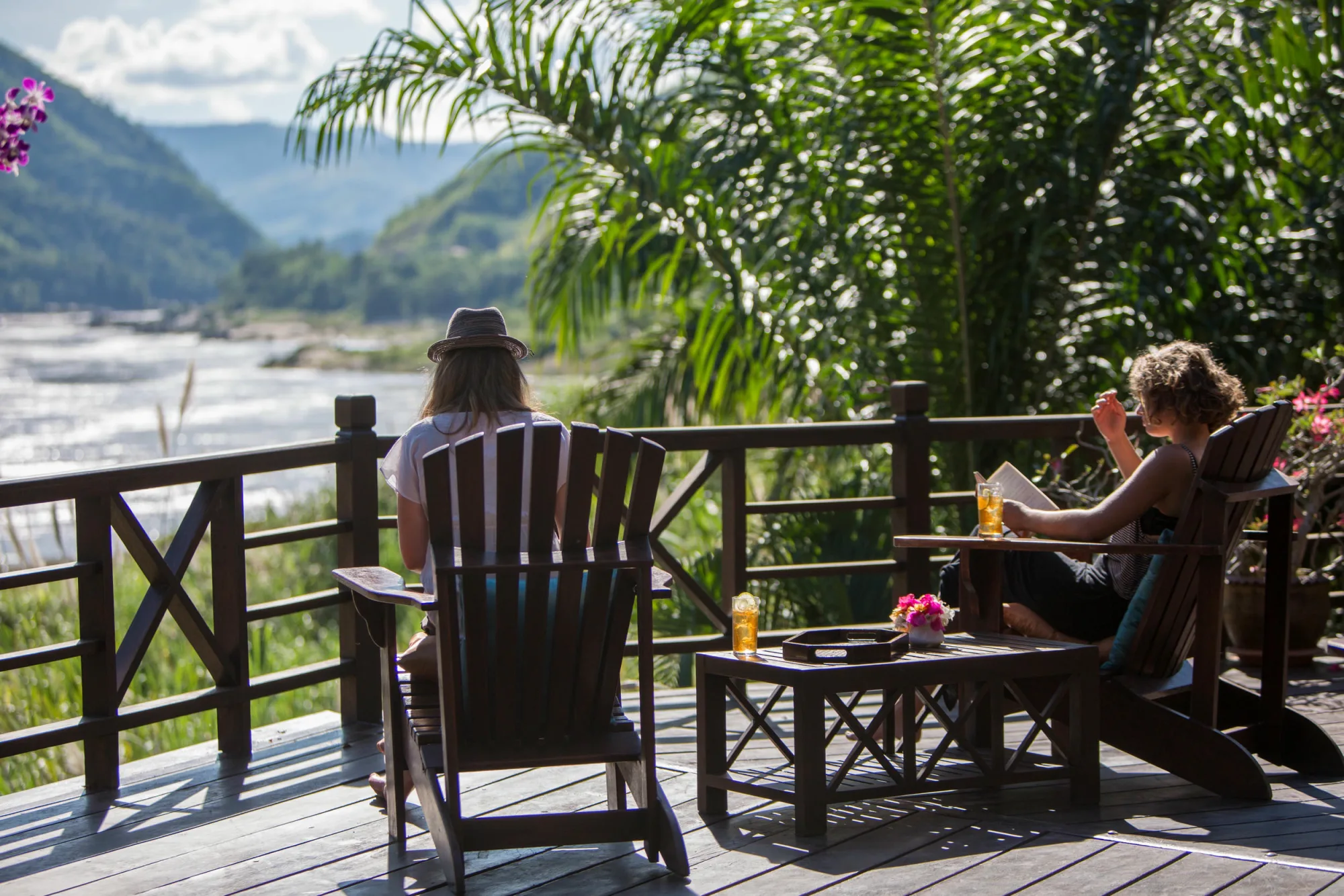 Guests relaxing in wooden armchairs on the terrace above the Mekong, with drinks and a book