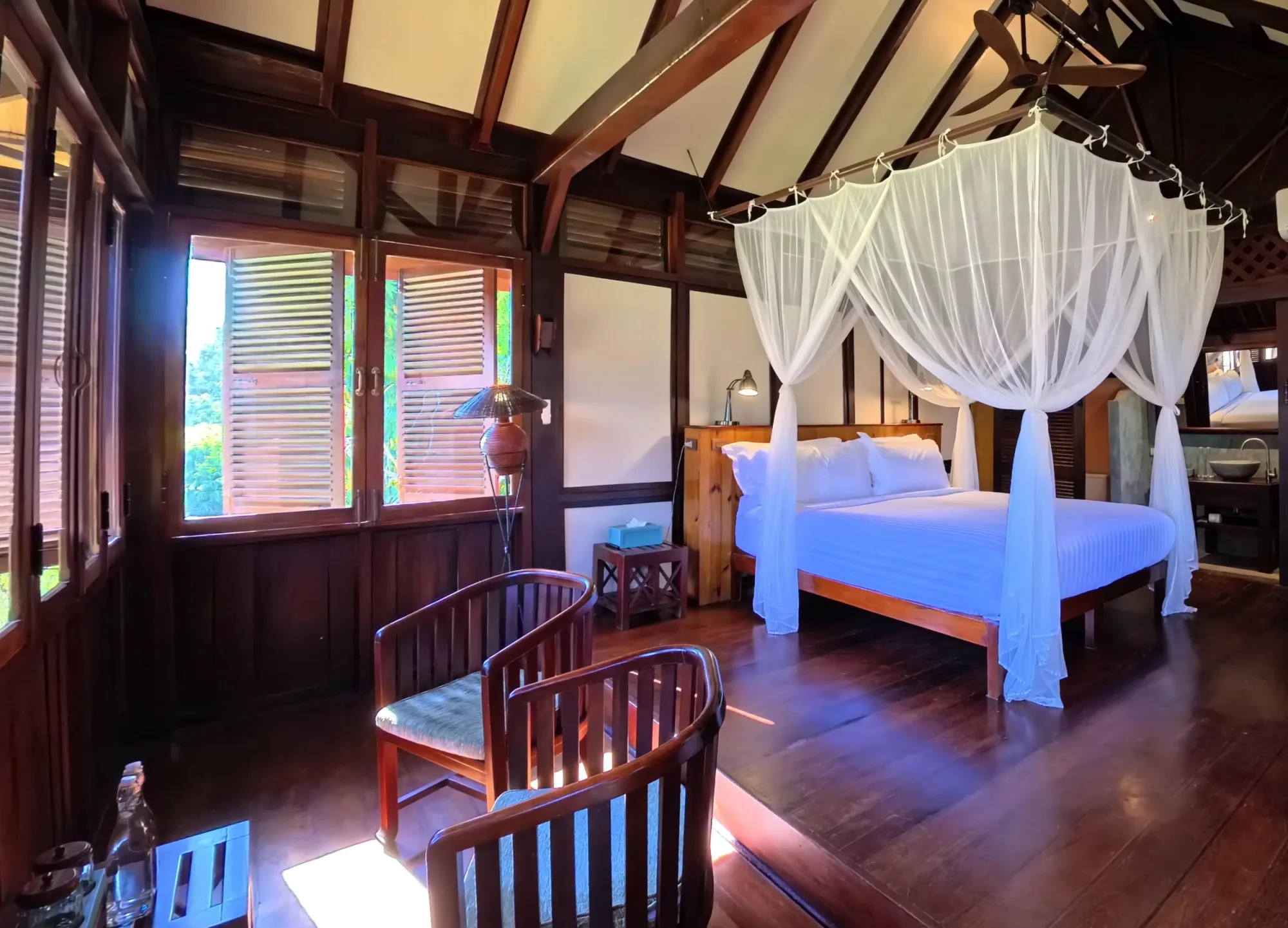 Bungalow interior with a king bed, mosquito net, wooden armchairs and shuttered windows