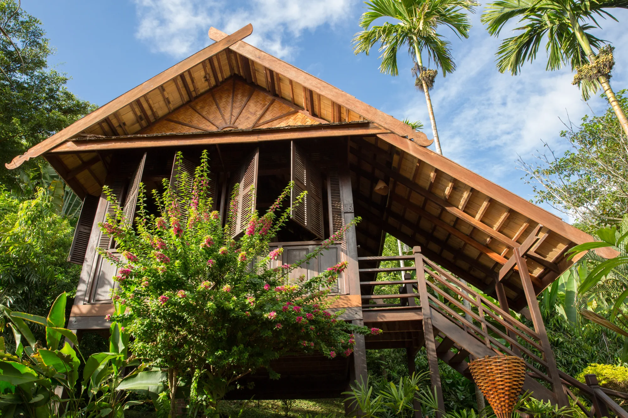 Teak bungalow with a peaked roof, open wooden shutters and a stairway, framed by bougainvillea and palms