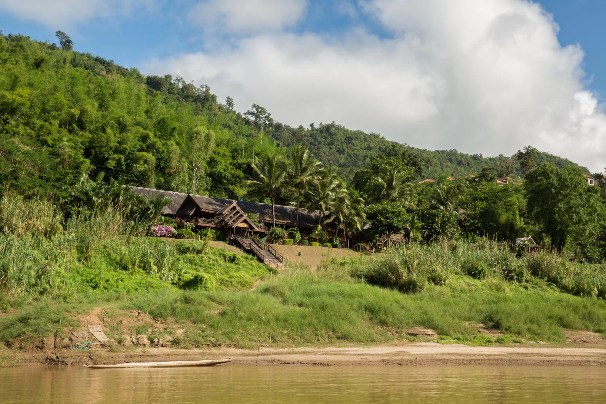 The long shingle roofs of Luang Say Lodge seen from the river, set among palms on the bank