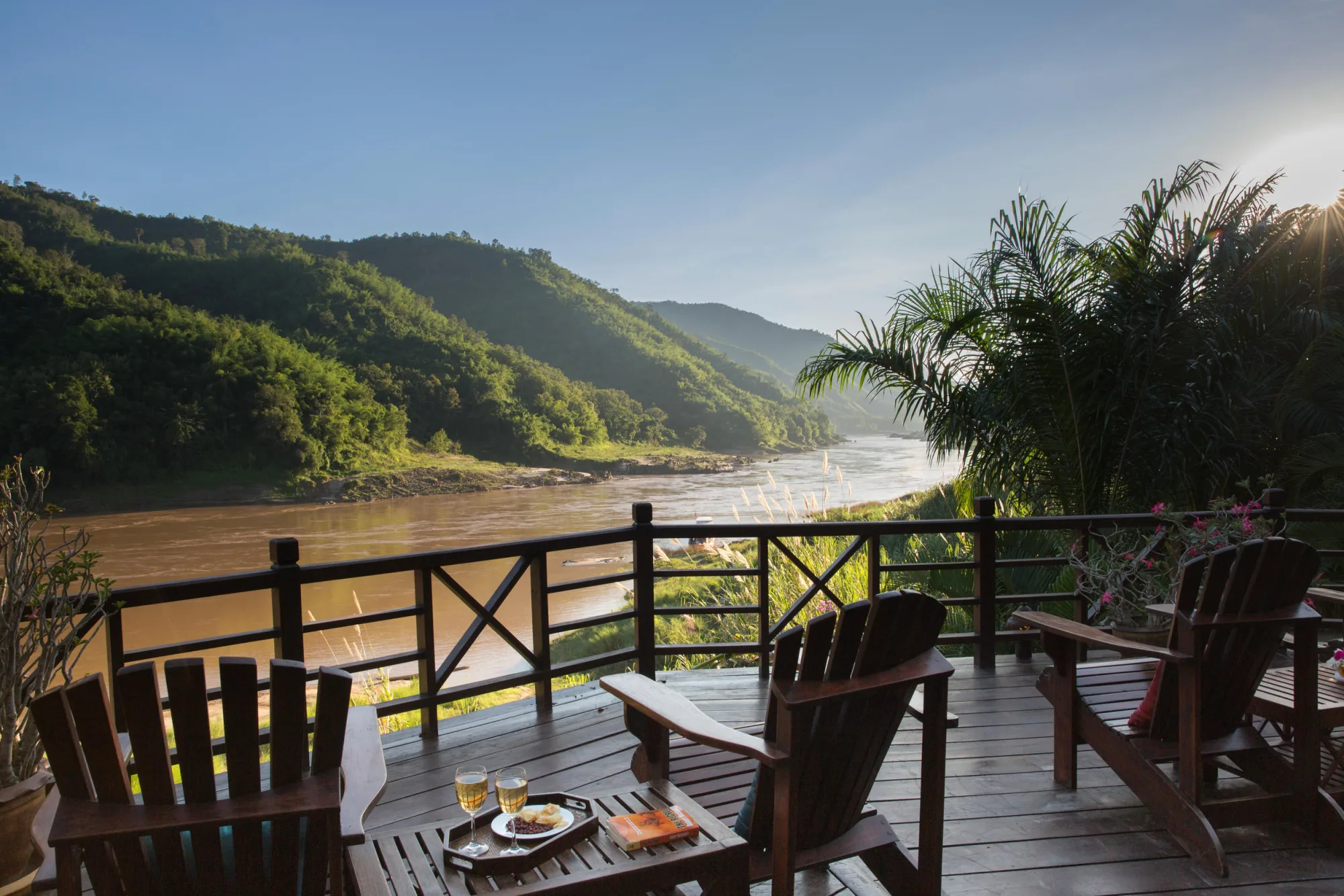 Wooden deck chairs on a riverside terrace overlooking the Mekong River and forested hills in late-afternoon light
